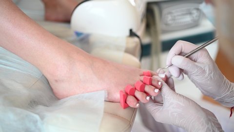 Pedicurist doing white nail polish on client legs using shellac lamp and toe finger separator. Professional medical pedicure procedure. Foot treatment in SPA salon. Podiatry clinic. Hands in gloves