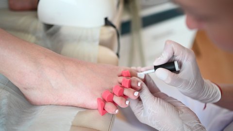 Pedicurist doing white nail polish on client legs using shellac lamp and toe finger separator. Professional medical pedicure procedure. Foot treatment in SPA salon. Podiatry clinic. Hands in gloves