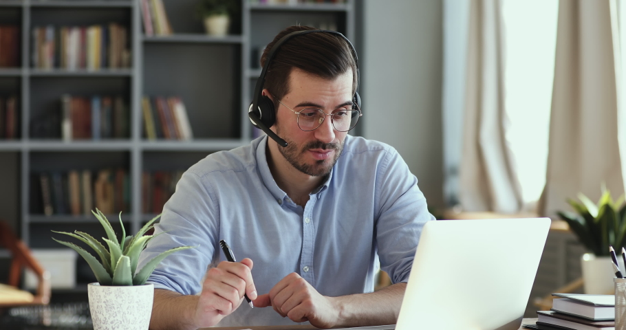 Confident young handsome businessman in headphones with mic holding video call with clients or partners, discussing project, writing down notes. Happy man listening webinar, studying online at home. Royalty-Free Stock Footage #1054153769