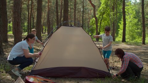 Two women and two boys having summer camping vacation in forest. Happy family of two mothers and two sons put up tent for camping. Slow motion, steadicam shot.