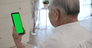 Over shoulder closeup view of senior elderly man holding smart phone vertical screen with green screen watching mobile video calling online to friend or family in virtual app.