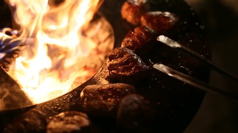 The chef roasts the meat on a metal hot surface. Meat steak is being prepared on a round steel iron outdoor grill with a cooktop and an open fire in the middle.