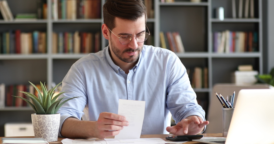 Focused young businessman in glasses holding paper receipt, doing accounting at office. Concentrated bearded man calculating household bills, managing monthly incomes and outcomes alone at home. | Shutterstock HD Video #1054161494