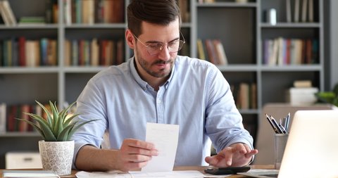 Focused young businessman in glasses holding paper receipt, doing accounting at office. Concentrated bearded man calculating household bills, managing monthly incomes and outcomes alone at home.