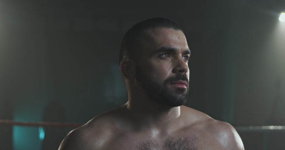 Portrait shot of handsome brutal Caucasian young man with beard turning face to camera and looking in dark boxing ring. Close up of sporty sweaty male boxer with serious face in darkness. Royalty-Free Stock Footage #1054165640