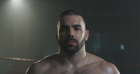Portrait shot of handsome brutal Caucasian young man with beard turning face to camera and looking in dark boxing ring. Close up of sporty sweaty male boxer with serious face in darkness.