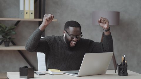 Overjoyed black male student sit at desk look at laptop screen scream passing exam getting high grade, excited african American guy feel euphoric receiving pleasant email message on computer