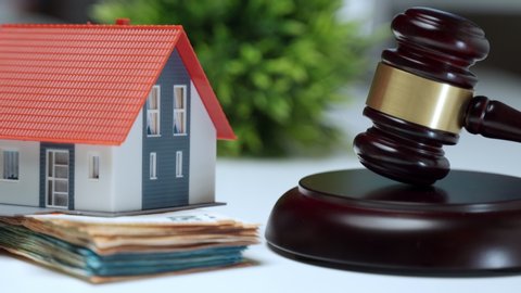 Miniature plastic house staying on stack of money near the wooden judge gavel, property auction concept. Financial transactions, investment and expenses during buying or selling house process