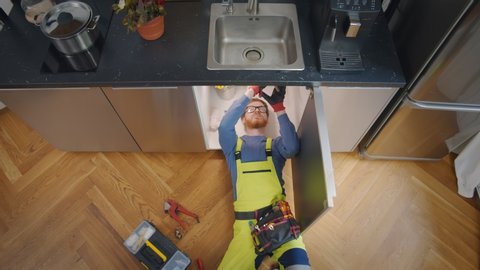 Top view of repairman in uniform lying under sink in kitchen fixing or cleaning pipes. Handyman repairing leaking faucet in home kitchen