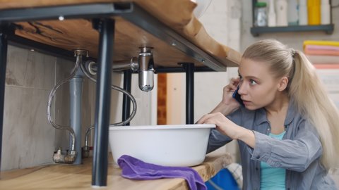 Young Woman Calling Plumber Crouching with Basin In Front Of Water Leaking From Sink Pipe Sitting on Bathroom floor. Housewife calling for plumber