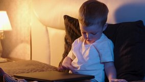 Cute caucasian boy in bedroom late at night sitting on bed opening laptop, typing something interesting surfing Internet. night light in room, moonlight shines from window. Dolly slides through video.