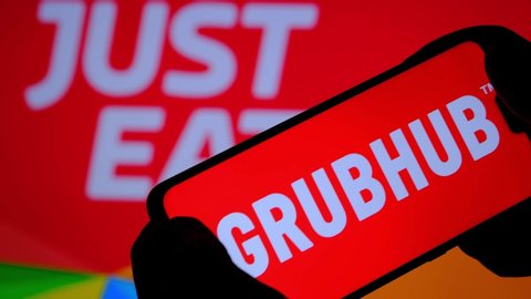 Stone / United Kingdom - June 11 2020: GRUBHUB company logo seen on smartphone silhouette and JUST EAT food delivery logo on the blurred background. Concept for company merger deal.