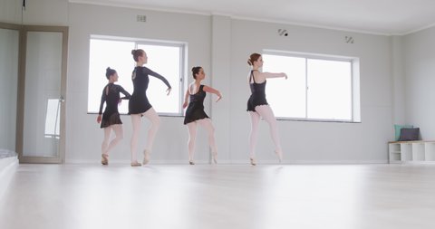 Four Caucasian female ballet dancers wearing black tricots practicing a dance routine during a ballet class in a bright studio, in slow motion.