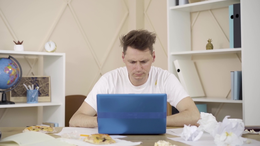 Stressed brunette Caucasian man holding head with hands and sighing. Portrait of young tired male freelancer working online in messy room. Lifestyle, freelance, remote work. | Shutterstock HD Video #1054179560