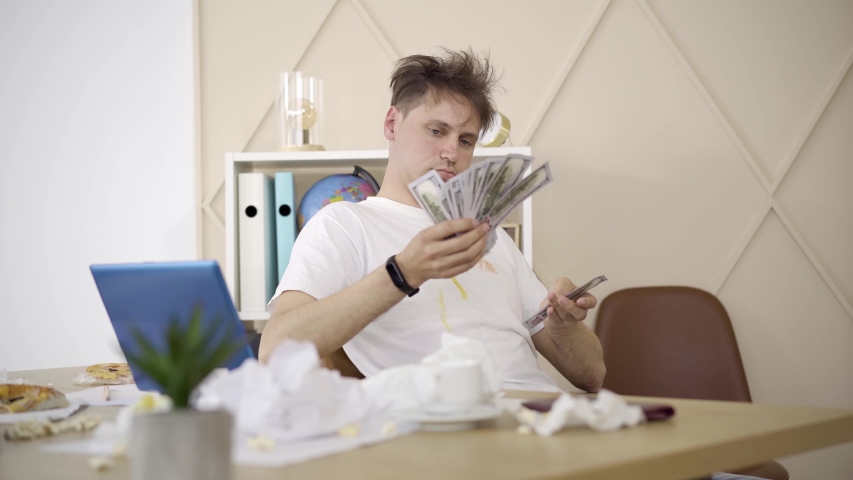 Portrait of young man counting money as sitting at the table in messy room. Brunette Caucasian guy in dirty T-shirt having good income working from home. Remote work, freelance lifestyle. Royalty-Free Stock Footage #1054179650