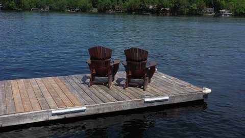 Two chairs on a wooden dock facing a calm lake in Ontario, Canada. Drone footage circling around the chairs