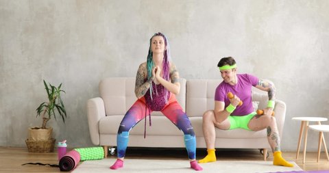 Funny man and woman newcomers in sport are doing fitness exercises at home. Girl doing squats, man raising dumbbells sitting on sofa. Joke, mem, humor, parody. Fitness, workout, training concept.