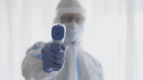 A male doctor or healthcare worker wearing a personal protective kit pointing digital infrared forehead thermometer or temperature gun in the hospital or clinic amid Coronavirus or COVID 19 epidemic