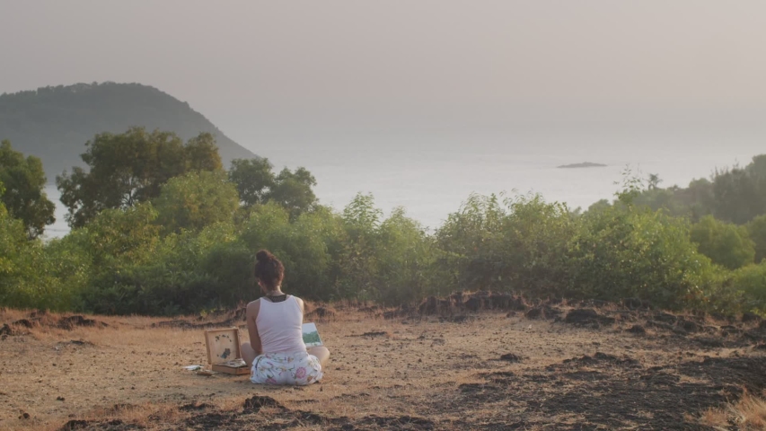Back view of young painter sitting outdoors in mountains painting marine landscape. Unrecognizable female artist working on fresh air in morning light slow motion. Summer vacation lifestyle relaxation