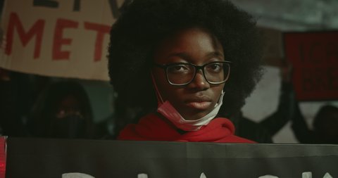 Close up view of african american millennial girl holding carton placard protesting against police brutality and racism. People with banners chanting at background. Concept of riot
