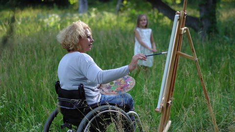 The artist in a wheelchair resorts a portrait of a little girl in nature. The young model is tired and trying to move. An artist with an easel in nature. Prores 422.