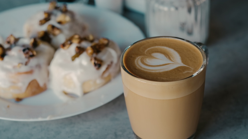 Lovely latte leaf design in glass cup next to fresh icing buns serving cappuccino woman hand taking it slow motion closeup with 4k. Restaurant food lifestyle
 | Shutterstock HD Video #1054195622