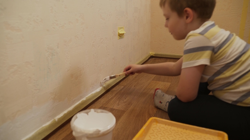 Caucasian boy sits on the floor and gently paints a wall with white paint with a brush near the skirting board and masking tape