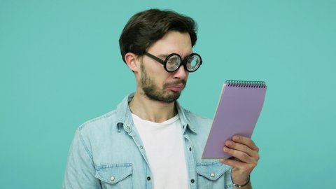 Comical weird geek in glasses studying. Clever bearded guy with round diopter eyeglasses reading notebook entries and gazing at camera, checking list-to-do. studio shot isolated on blue background