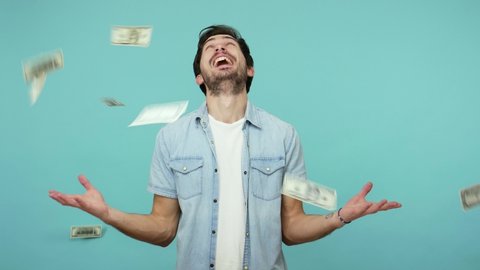 Excited happy lottery winner, glad bearded guy in jeans shirt looking up and shouting from happiness while money rain falling from above, rich man enjoying wealth. indoor studio shot, blue background