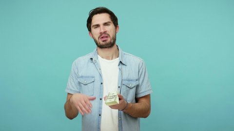 Handsome bearded guy in jeans shirt scattering dollars with arrogant haughty expression, throwing around cash, squandering, wasting money carelessly. indoor studio shot isolated on blue background