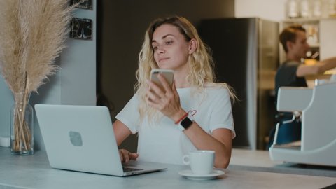 Blonde caucasian woman  talking by phone during working with laptop in coffee shop with barista on background. Female freelancer communicates with someone and typing on computer. 
