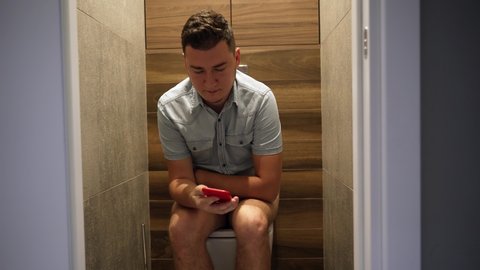 A young man sits on a toilet in a restroom and looks at a smartphone. He is surprised by something