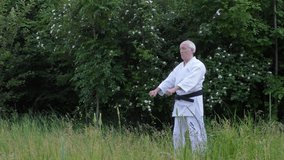With a black belt, an old athlete is training formal karate exercises on a background of nature