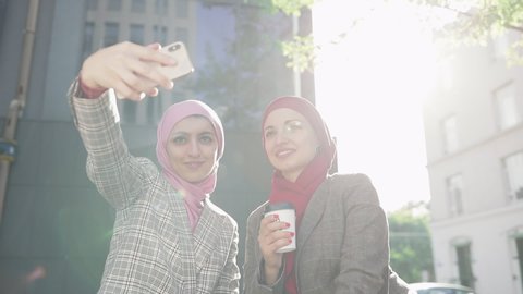 Two young Muslim women wearing hijab headscarf making selfie at phone, sitting together at bench laughing smile to eash other, modern lifestyle, technology, smartphone usage, leisure time.