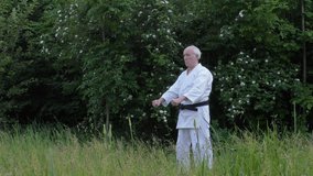 With a black belt, an old athlete does formal karate exercises on a background of nature