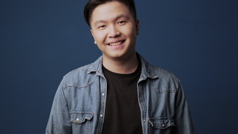 A happy young asian man wearing casual clothes is smiling and looking to the camera standing isolated over dark blue wall background in studio