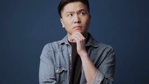 A serious young asian man wearing casual clothes is thinking about something standing isolated over dark blue wall background in studio