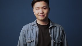 A successful young asian man wearing casual clothes is laughing and looking to the camera standing isolated over dark blue wall background in studio