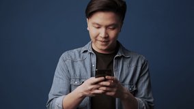 A happy smiling young asian man is using his smartphone isolated over dark blue wall background in studio