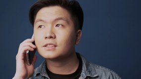 A close-up view of an attractive young asian man is talking on the phone isolated over dark blue wall background in studio