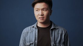 A displeased young asian man wearing casual clothes covers nose with hand and showing stop gesture isolated over dark blue wall background in studio