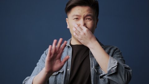 A displeased young asian man wearing casual clothes covers nose with hand and showing stop gesture isolated over dark blue wall background in studio