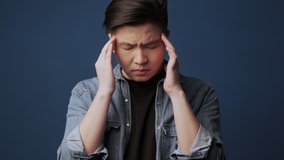 A young asian man with closed-eyes wearing casual clothes is massaging his temples isolated over dark blue wall background in studio