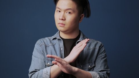 A serious young asian man wearing casual clothes saying NO while crossing his hands standing isolated over dark blue wall background in studio
