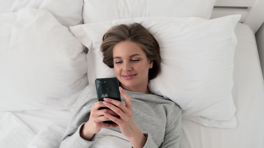 A happy smiling young woman is watching something good on her smartphone while lying in the white bed at home