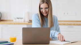 A lovely young blonde woman wearing glasses is writing something looking at her laptop sitting in the white kitchen at home