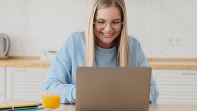 A happy satisfied young blonde woman wearing glasses is using her laptop working in the white kitchen at home
