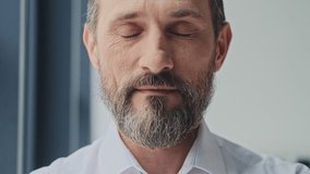 A close-up view of a calm mature man is opening his eyes and looking to the camera while standing in the office