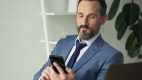 A successful smiling mature man is using his smartphone sitting in the office