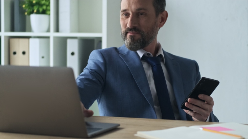 A confident smiling mature man is using his smartphone while working in the office Royalty-Free Stock Footage #1054208960